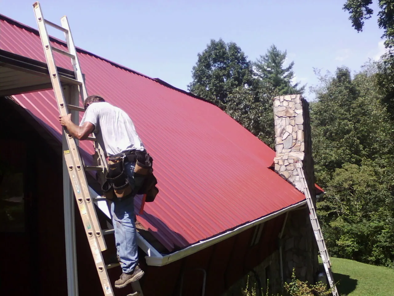 Roofer on a ladder beside a red metal roof and stone chimney — Soffit & Fascia Repair by Roofing Brothers in Wantagh