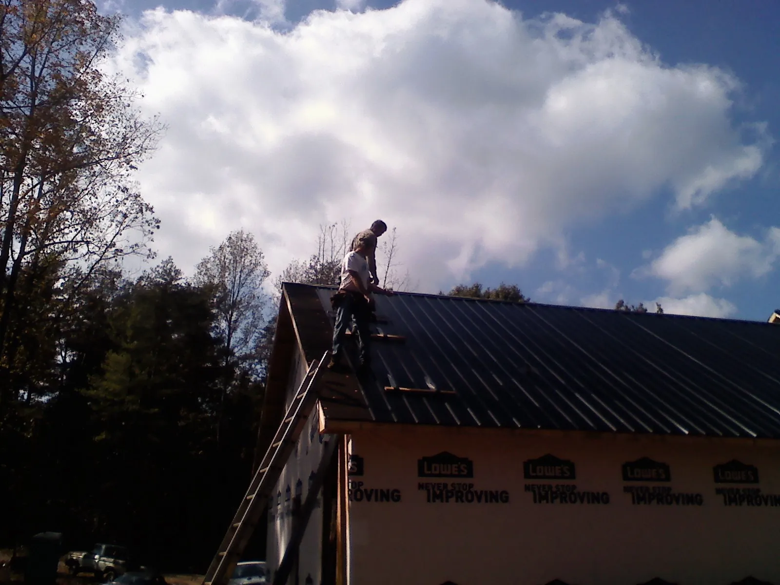 Roofer fastening dark metal panels high against a cloudy sky, from a Roof Inspection job in Wantagh